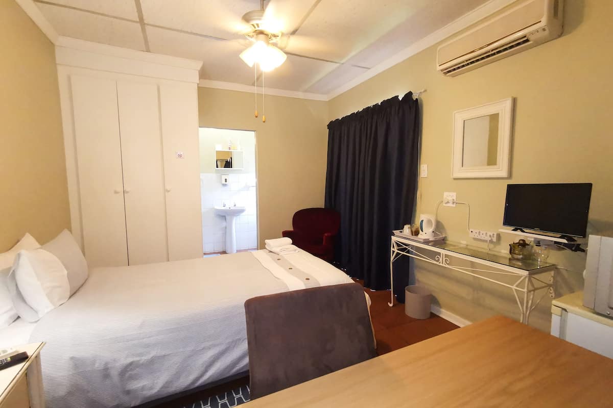 A hotel room with a bed, a desk, a chair, a TV, a ceiling fan, and a bathroom visible through an open door.