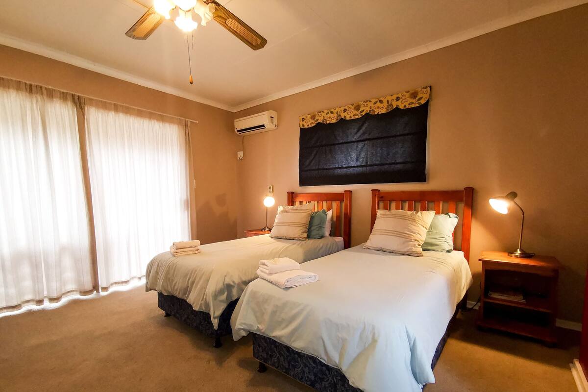 A hotel room with two beds, a ceiling fan, an air conditioning unit, a bedside lamp, and a large window with curtains.