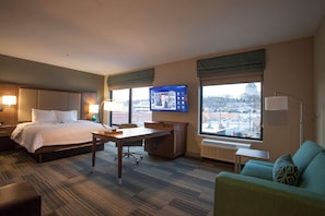 A hotel room with a bed, desk, and seating area, featuring a large window with a city view.