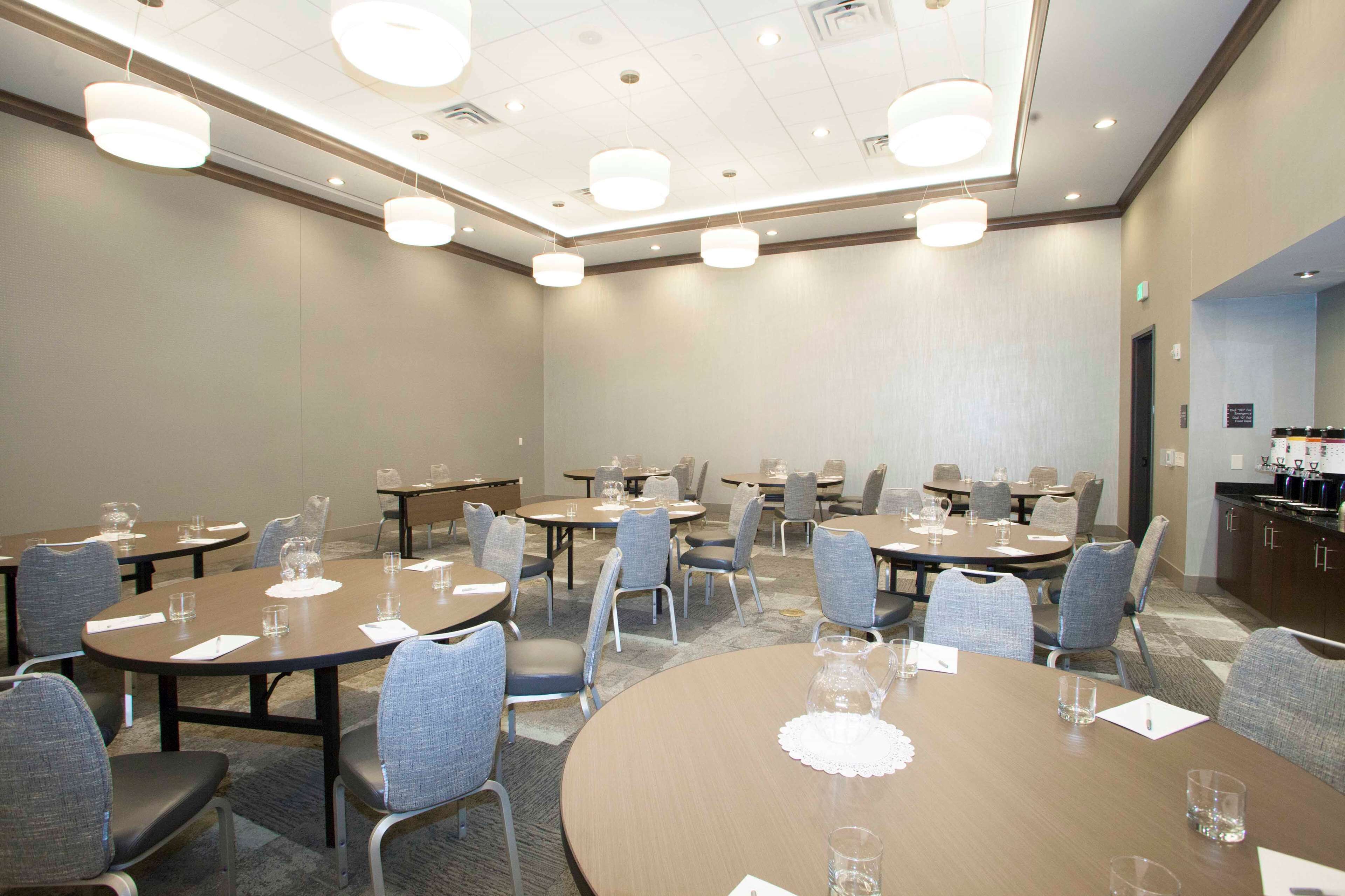 Een congreszaal met ronde tafels, stoelen en een kitchenette.