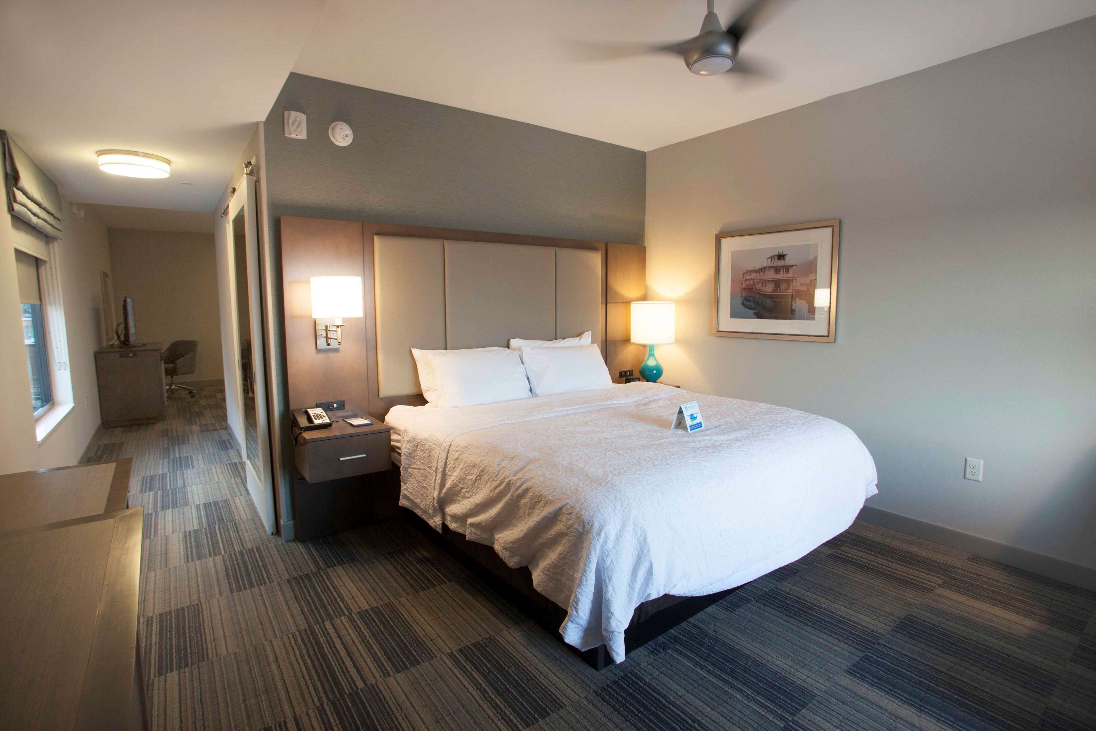 A hotel room with a large bed, a desk with a lamp, a phone, and a framed picture on the wall.