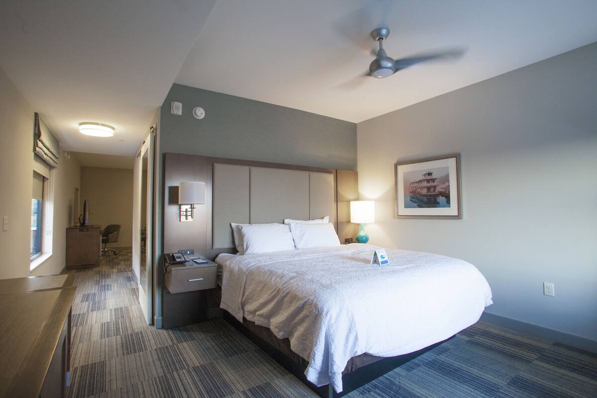 A hotel room with a large bed, a desk, a chair, a nightstand, a lamp, a ceiling fan, and a framed picture on the wall.