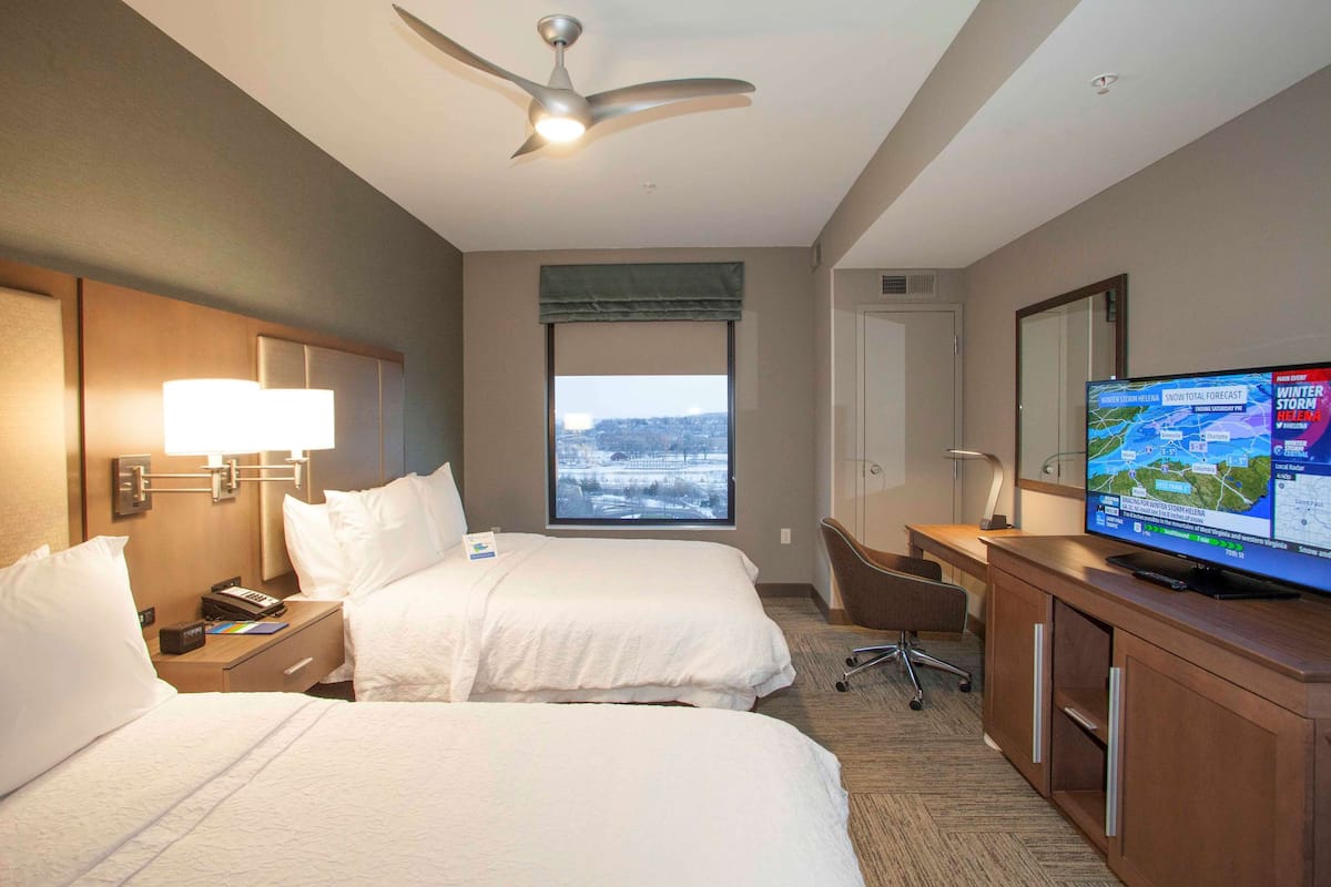 A hotel room with two beds, a desk, a television displaying a weather forecast, and a view of a snowy landscape through the window.