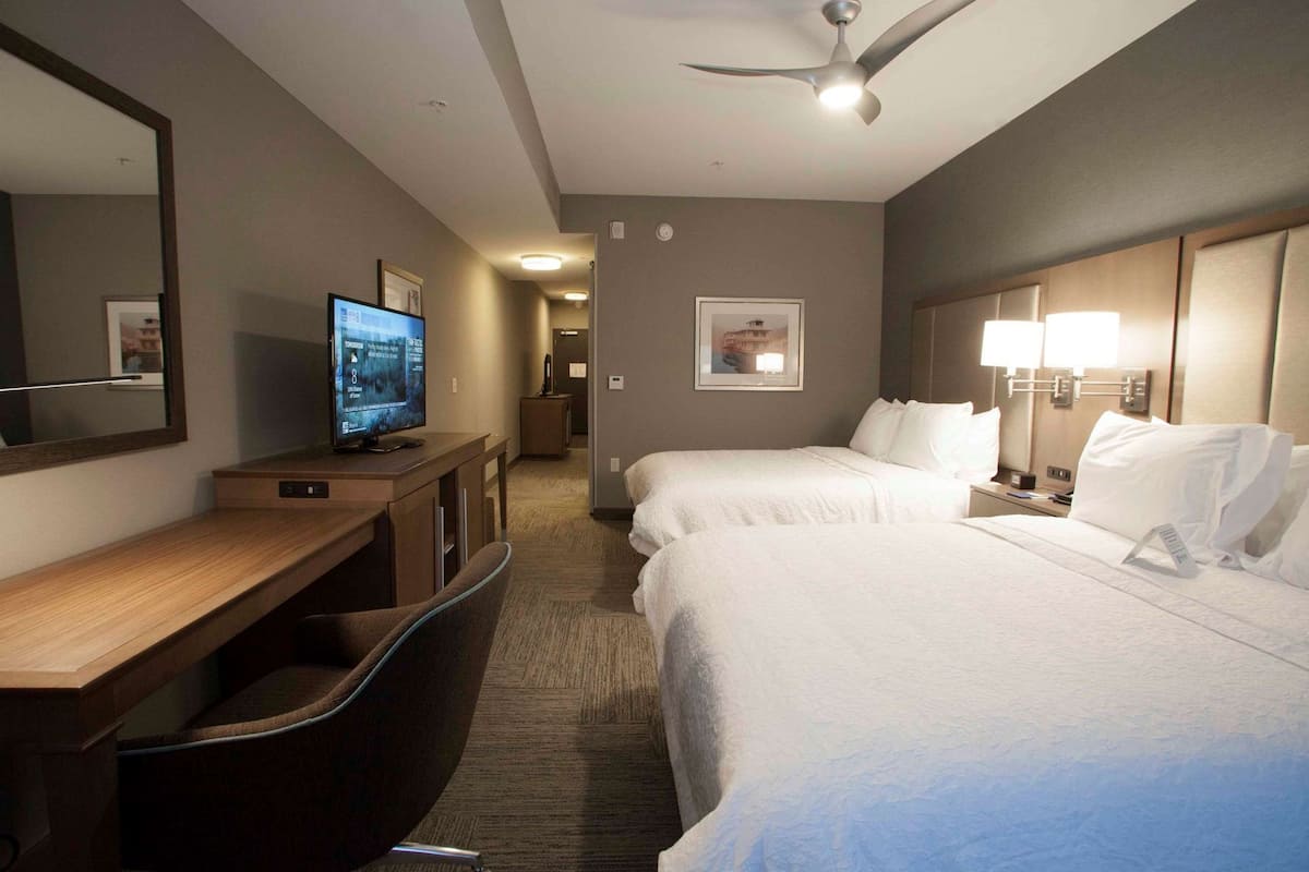 A hotel room with a desk, two beds, a television, a ceiling fan, and a hallway leading to another room.