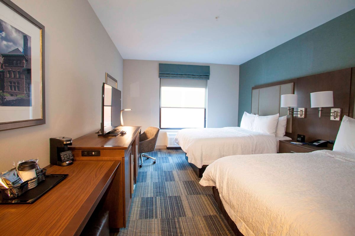 A hotel room with two beds, a desk with a coffee maker, a chair, and a large window.