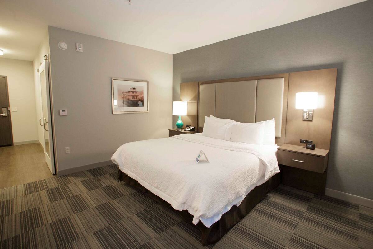 A hotel room with a large bed, a nightstand with a lamp, a wall-mounted clock, and a framed picture on the wall.