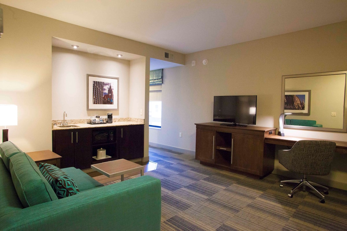 A hotel room with a green sofa, a small table, a TV, a desk, and a mirror.
