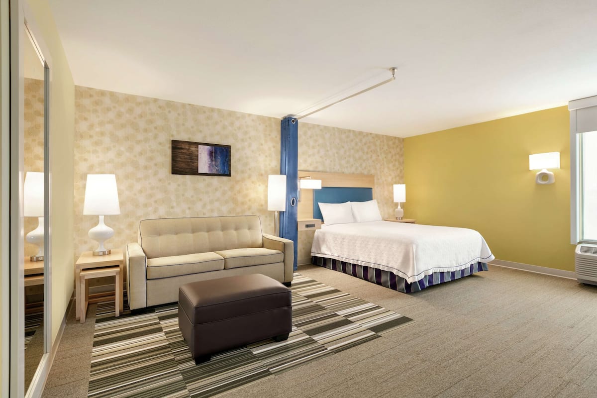 A hotel room with a sofa, a bed, a nightstand, a lamp, and a wall-mounted artwork.