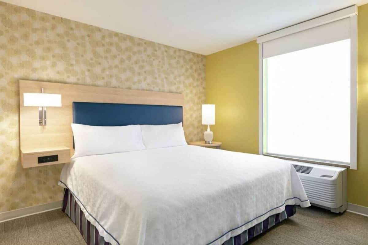 A hotel room with a bed, two bedside lamps, a window with a view, and a wall-mounted heater.