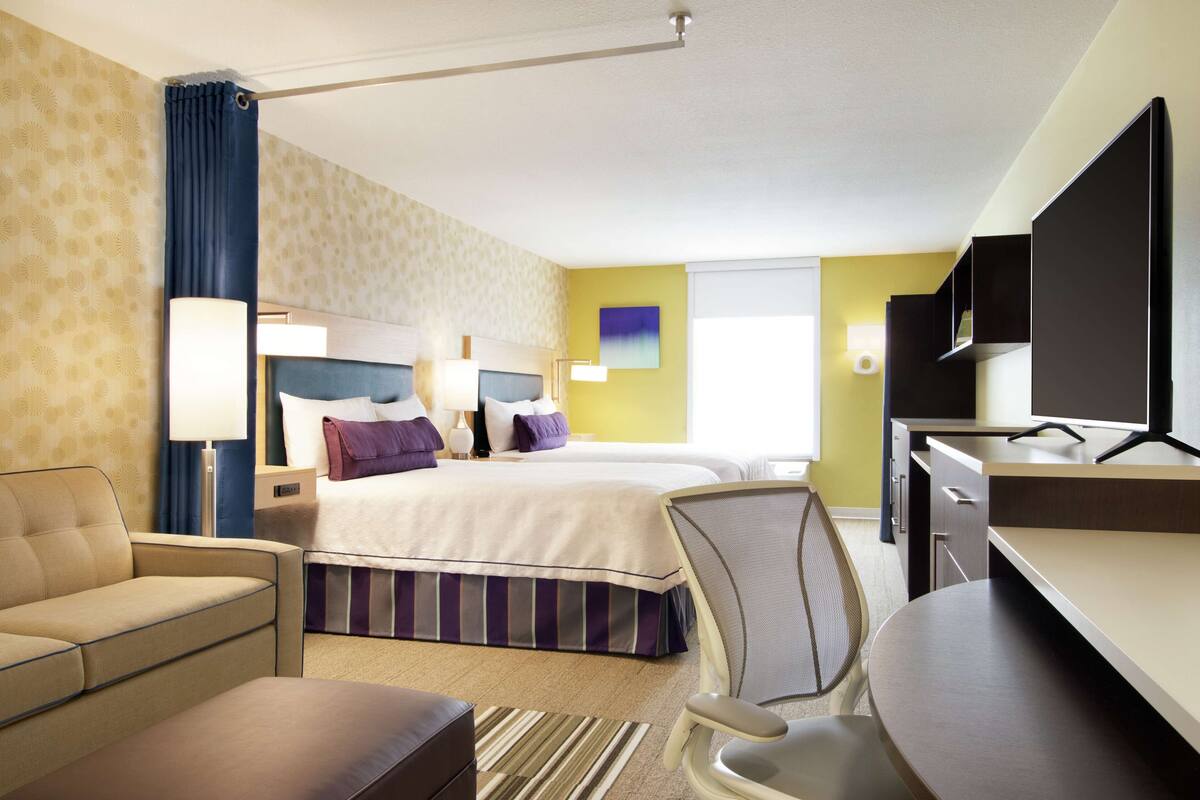 A hotel room with two beds, a desk, a chair, a sofa, and a TV.