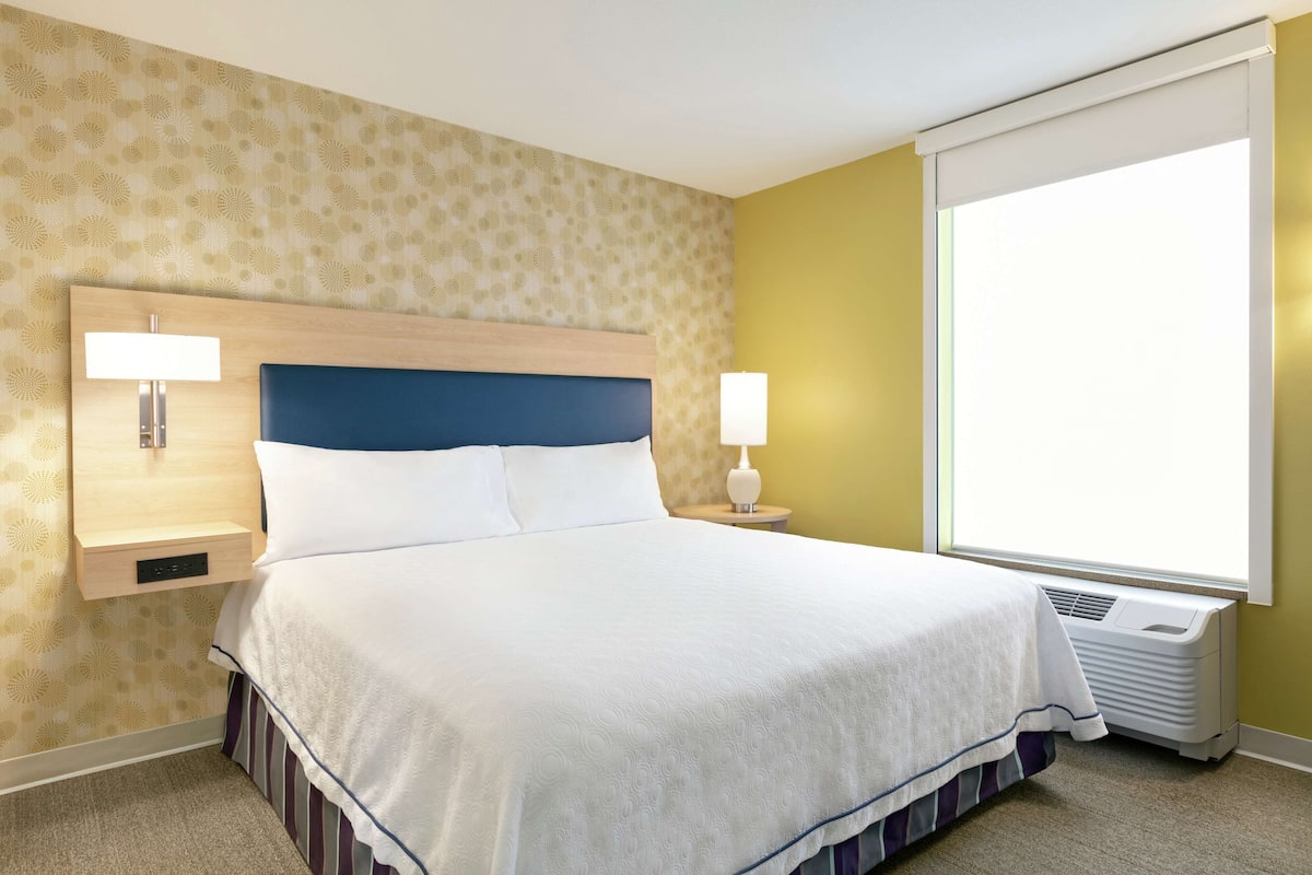 A hotel room with a large bed, a bedside table with a lamp, a wall-mounted heater, and a window with a view.