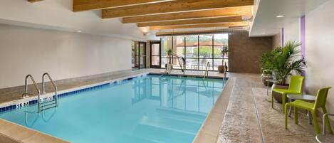 Una piscina interior con agua azul cristalina, vigas de madera en el techo y una zona de estar con sillas y mesas.