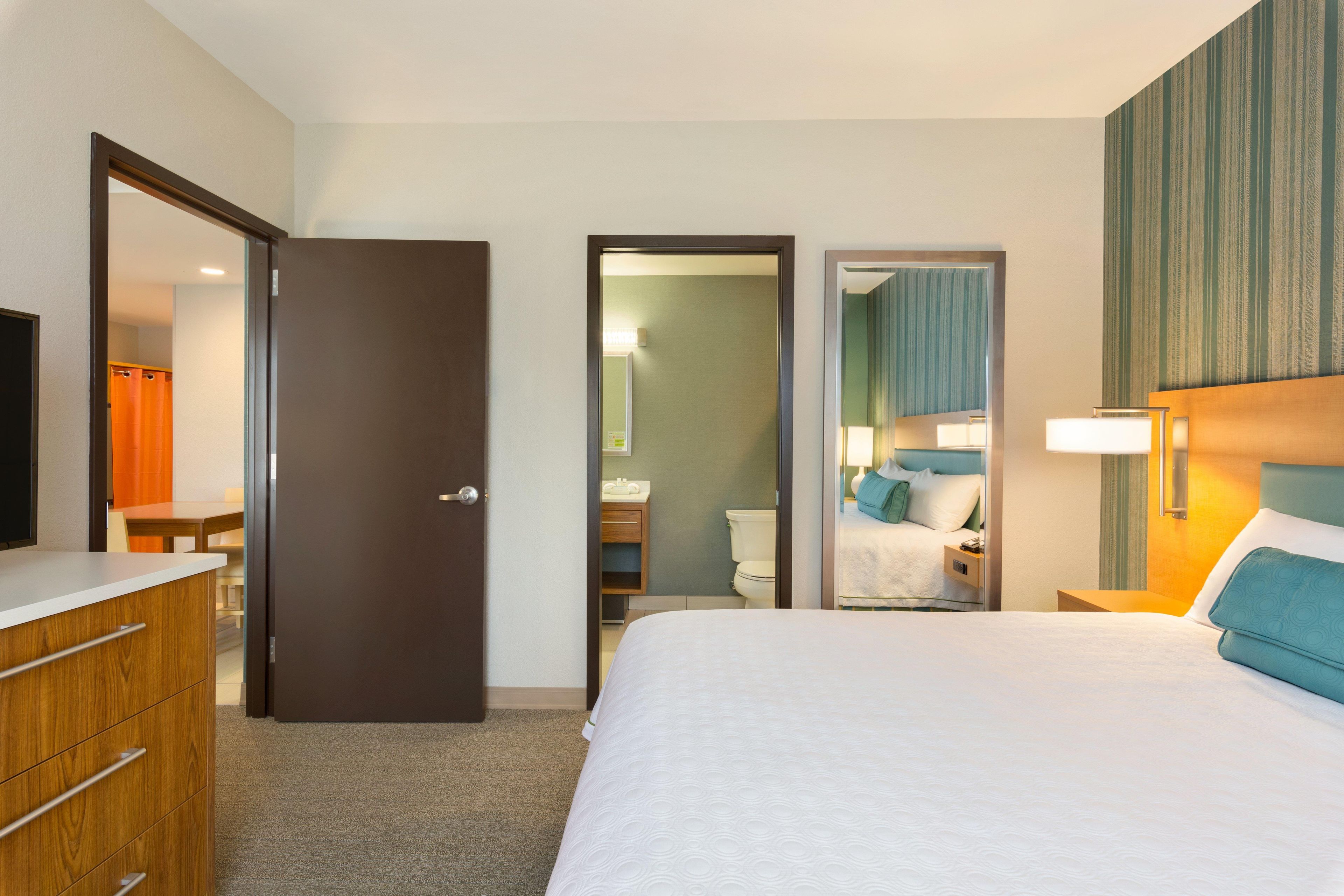 A hotel room with a bed, a dresser, a mirror, a bathroom with a toilet and sink, and a shower area.