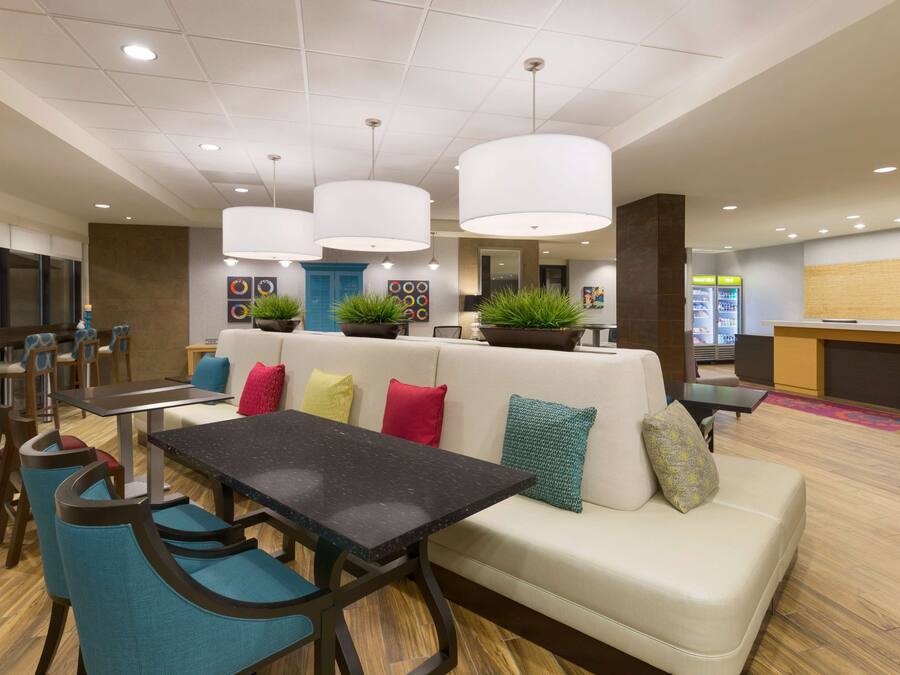 A modern lounge area with a black table, teal chairs, a white sofa with colorful cushions, and potted plants.