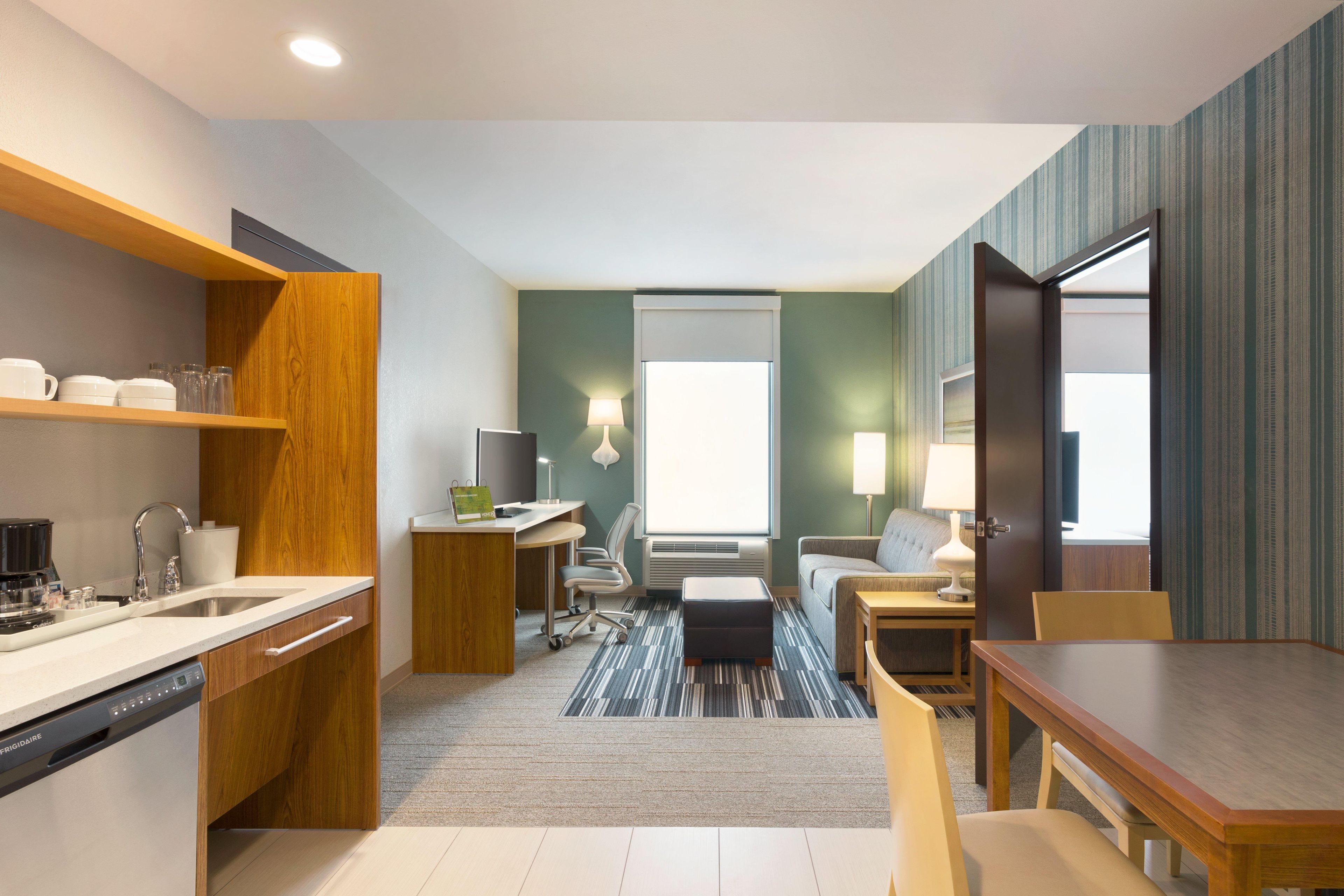 Habitación de hotel compacta con kitchenette, zona de estar con sofá y sillones, una pequeña mesa de comedor y ventana que proporciona luz natural.