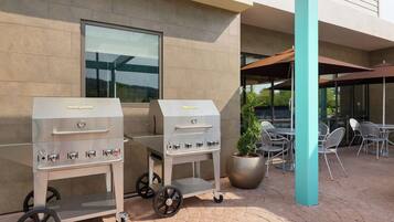 Two portable gas grills on a paved patio with outdoor seating and a building in the background.