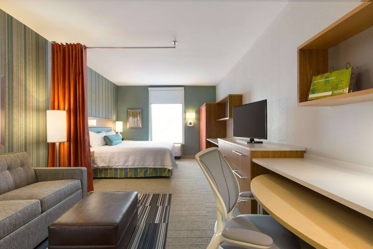 A hotel room with a bed, a sofa, a desk, a chair, a TV, and a shelf with a book and a bag.