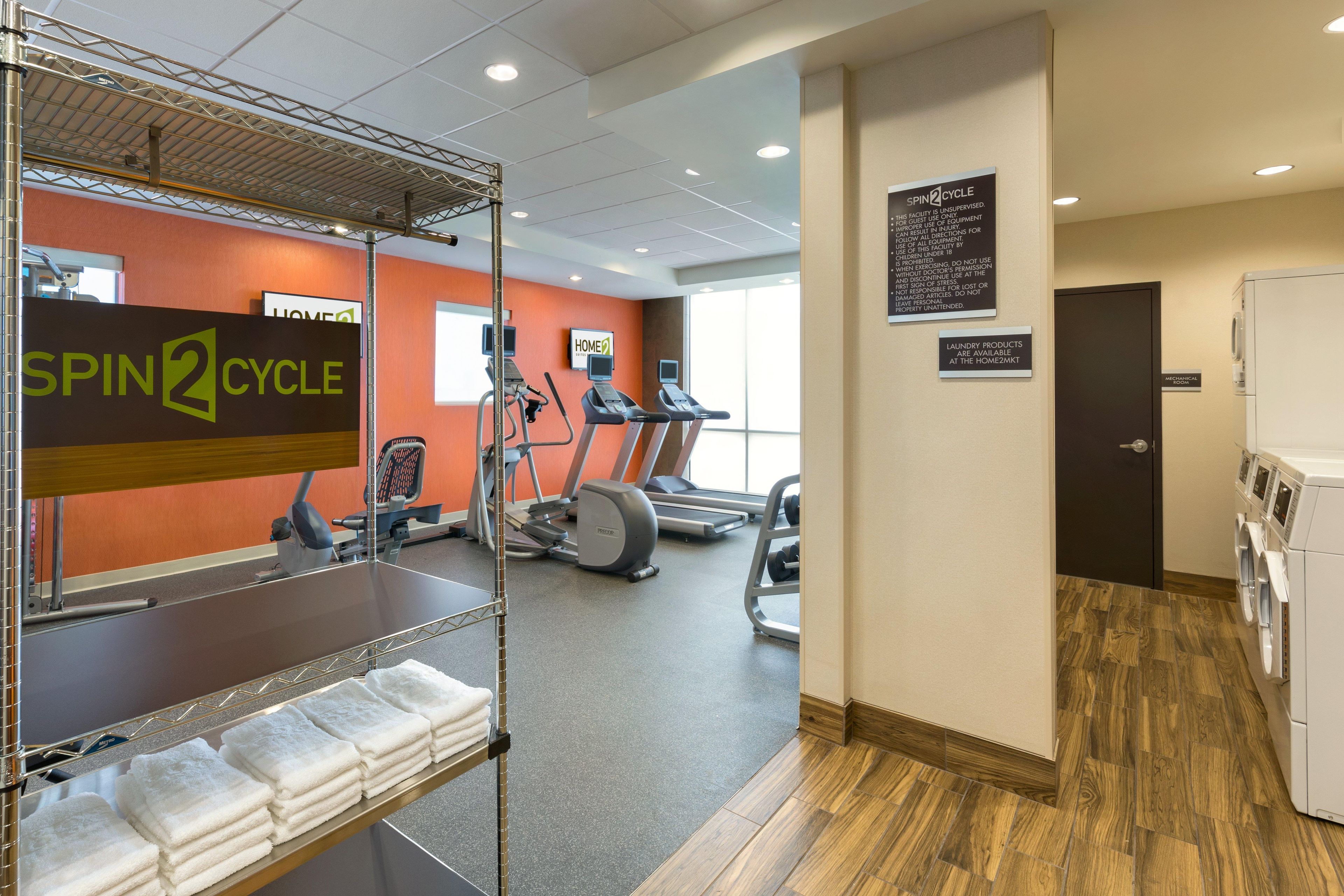 A gym with treadmills, elliptical machines, and a spin cycle area. There are towels on a rack, and a sign indicating laundry products are available.