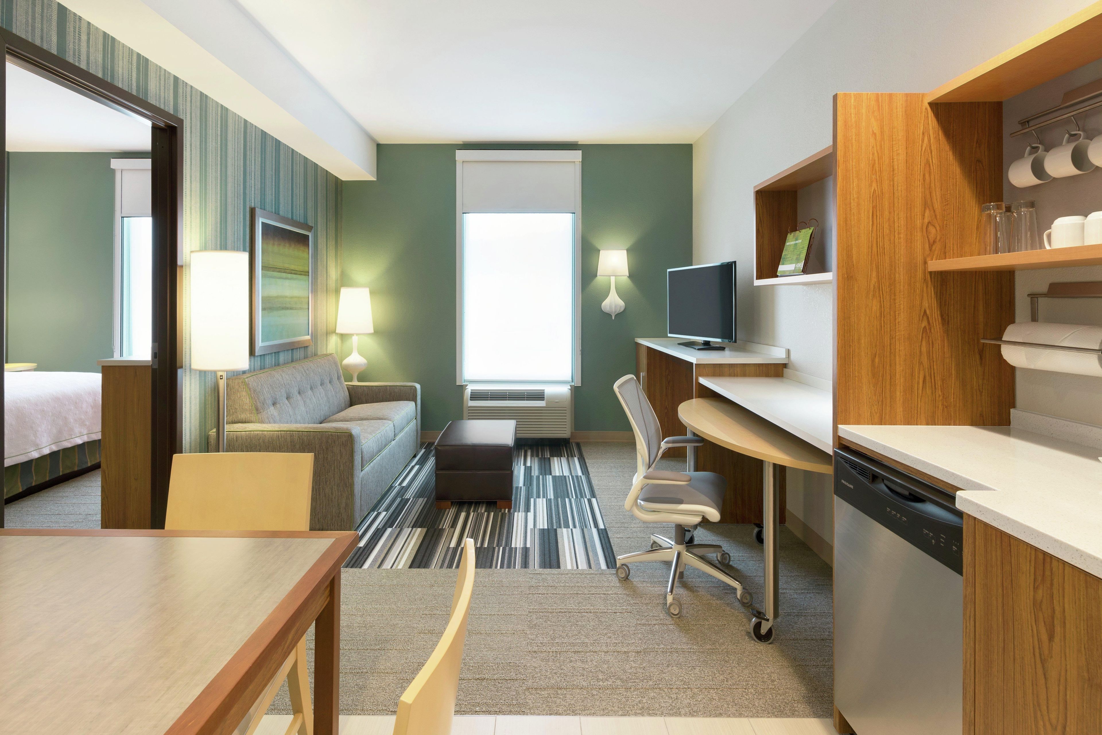 A hotel room with a bed, a sofa, a desk, a chair, a television, a lamp, and a kitchen area.
