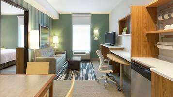 A hotel room with a bed, a sofa, a desk, a chair, a television, a lamp, and a kitchen area.