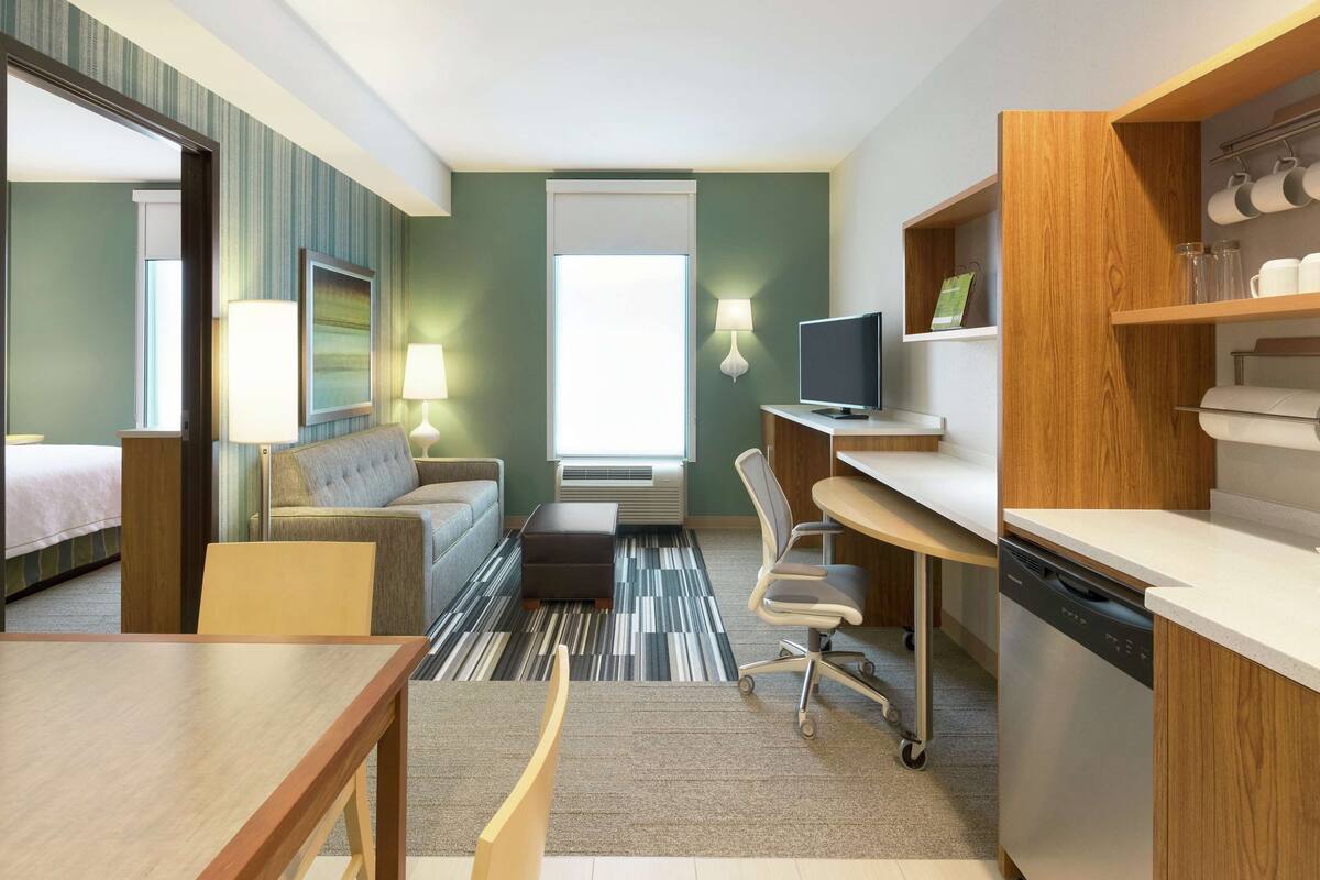 A hotel room with a bed, a sofa, a desk, a chair, a television, a dishwasher, and a window with blinds.
