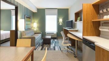 A hotel room with a bed, a sofa, a desk, a chair, a television, a lamp, and a kitchen area.