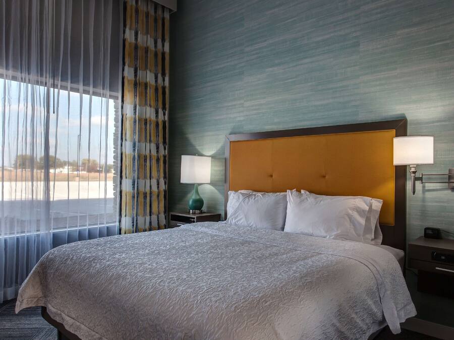 Habitación de hotel con una cama grande, dos lámparas de noche, una mesita de noche y vistas al exterior a través de cortinas traslúcidas.