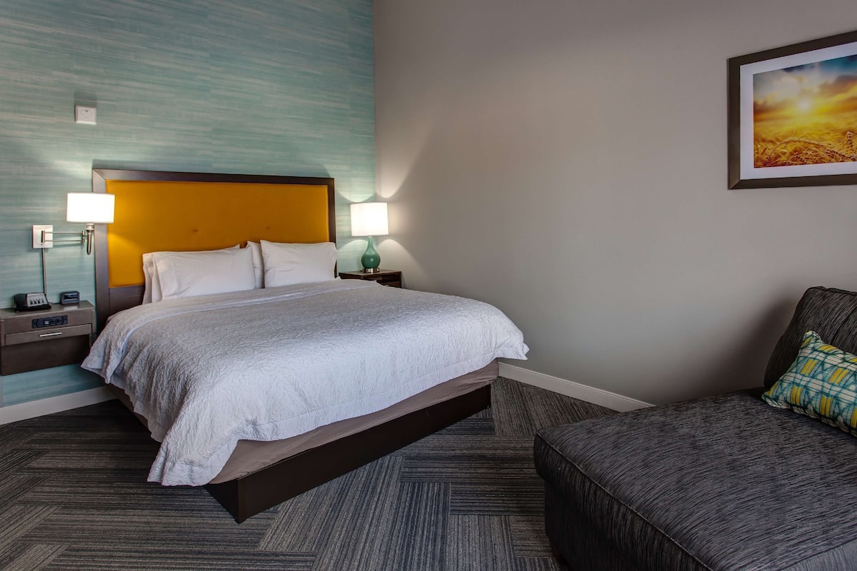 A hotel room with a bed, bedside tables, lamps, a chair, and a painting on the wall.