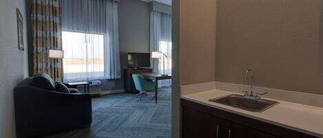 A hotel room with a kitchenette, a sofa, a desk, and a chair.