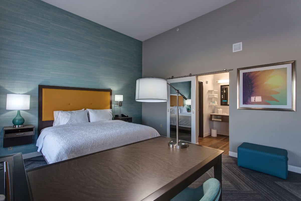 A hotel room with a bed, bedside tables, a desk, a chair, a lamp, a bench, and a bathroom visible through an open door.