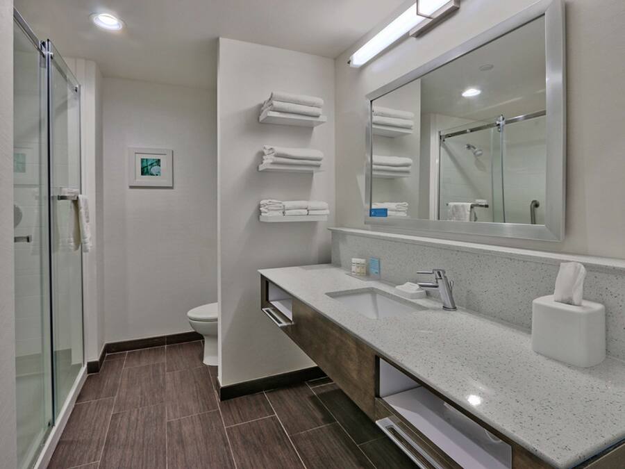 Un baño moderno con cabina de ducha de vidrio, un lavabo blanco con tocador de madera y un espejo grande sobre el lavabo.