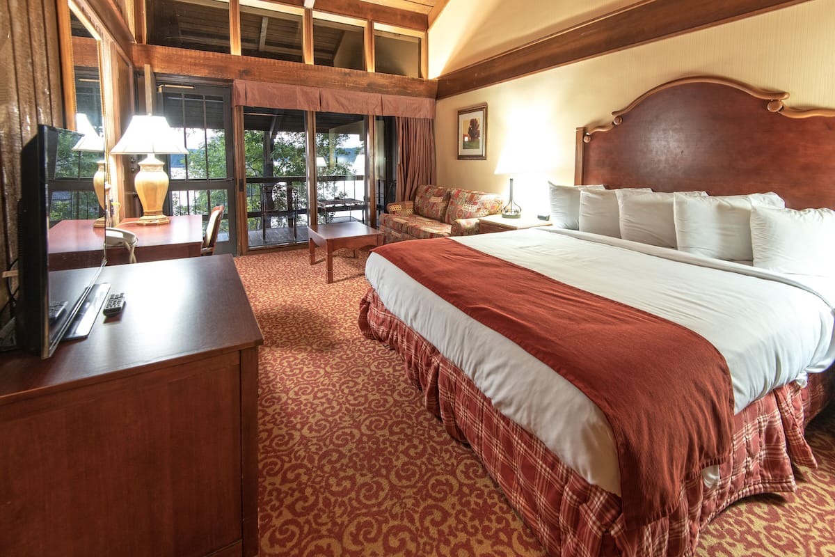 A hotel room with a large bed, a desk with a lamp, a chair, and a view of the outdoors through large windows.