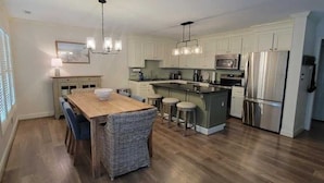 A modern kitchen with a central island, stainless steel appliances, and a dining area with a wooden table and wicker chairs. - Holden Haven 4 Bedroom Home by RedAwning (Supply)