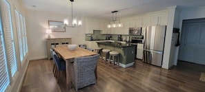 Cozinha moderna com ilha central, eletrodomésticos de aço inoxidável e área de jantar com mesa de madeira e cadeiras de vime.