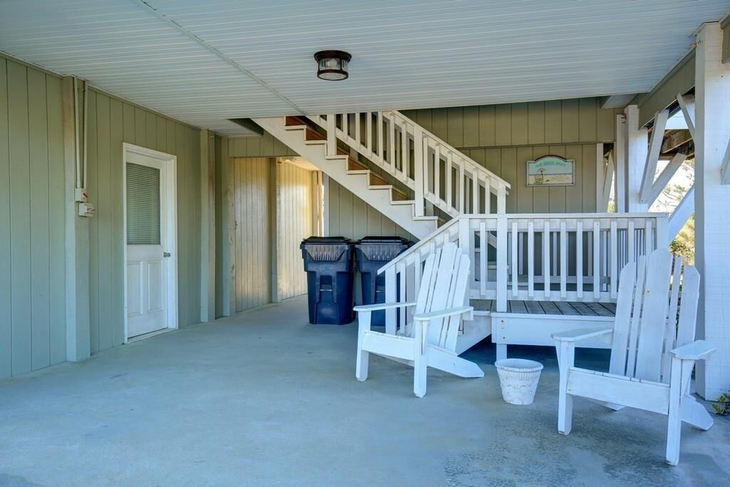 A covered porch with a staircase, white chairs, and trash bins.