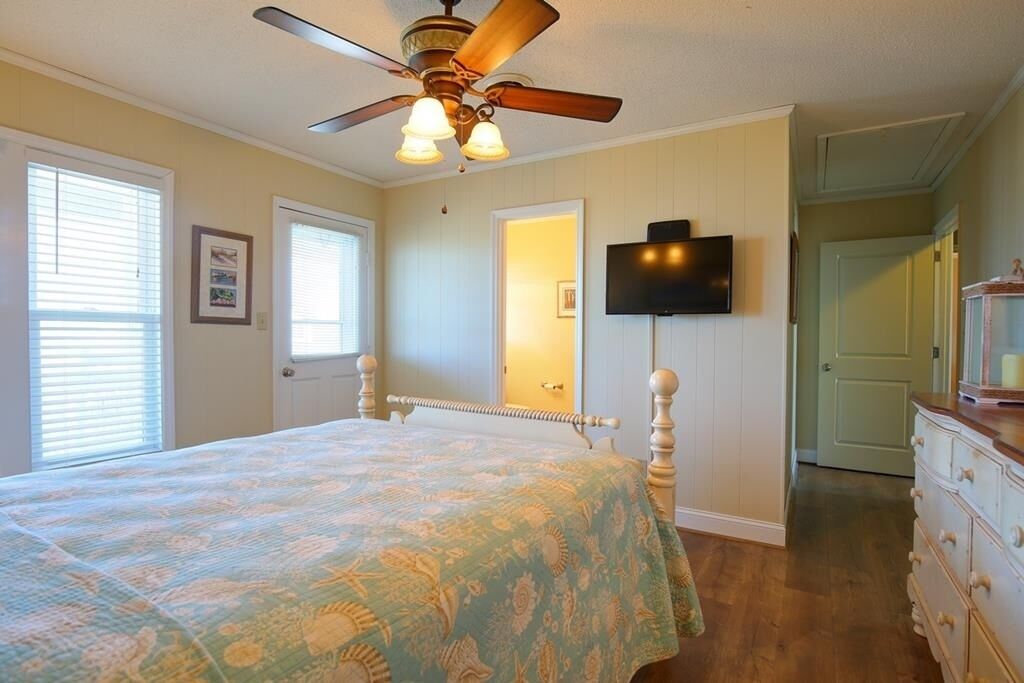 A bedroom with a bed, a ceiling fan, a television mounted on the wall, and a dresser.