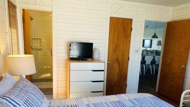 Un dormitorio con cama, televisor sobre un aparador de madera, baño con ducha, y un comedor visible a través de una puerta abierta.