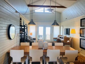 A dining area with a wooden table and white chairs, a ceiling fan, and a TV mounted on the wall.