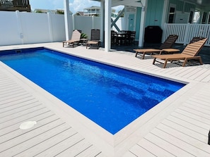Uma piscina retangular com um deck de madeira ao redor, uma área de varanda coberta com cadeiras e uma cerca branca.