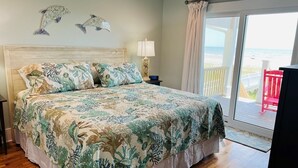 Une chambre à coucher avec un lit, une table de chevet, une lampe et une vue sur la plage, visible à travers une porte coulissante.
