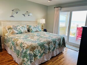 Quarto com cama, mesa de cabeceira, abajur e vista para a praia através de uma porta deslizante.
