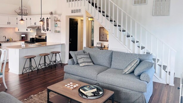 Un vaste espace de vie comprenant un canapé gris, une table basse avec des livres et un plateau, une cuisine avec des meubles blancs et un escalier avec des rampes blanches.