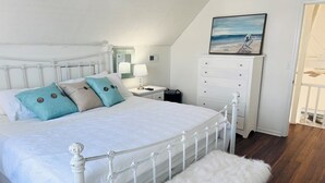 Une chambre à coucher avec un lit en métal blanc, un banc blanc, une table de chevet avec une lampe, une commode et un tableau représentant une scène de plage.