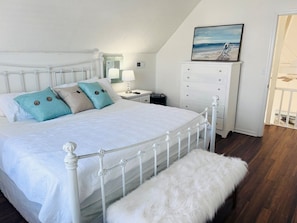 Quarto com estrutura de cama em metal branco, um banco branco, mesa de cabeceira com abajur, cômoda e uma pintura com cena de praia.