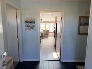 Um corredor dá para uma sala de estar, com ventilador de teto, geladeira e uma janela com persianas.