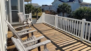 Una terraza de madera con barandas blancas, un espacio cubierto con una silla, y casas al fondo.
