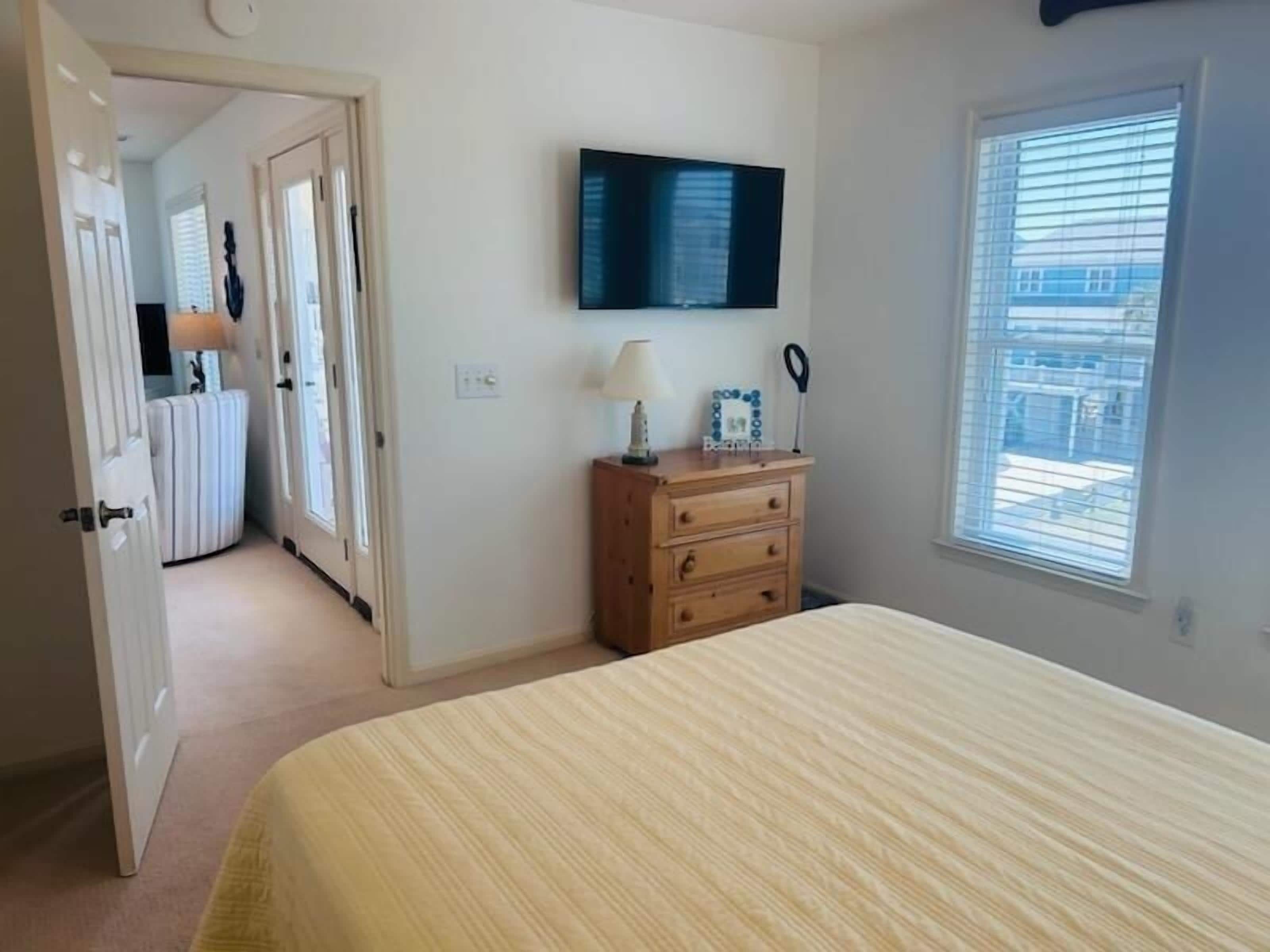 A bedroom with a bed, a dresser, a television mounted on the wall, and a window with blinds.
