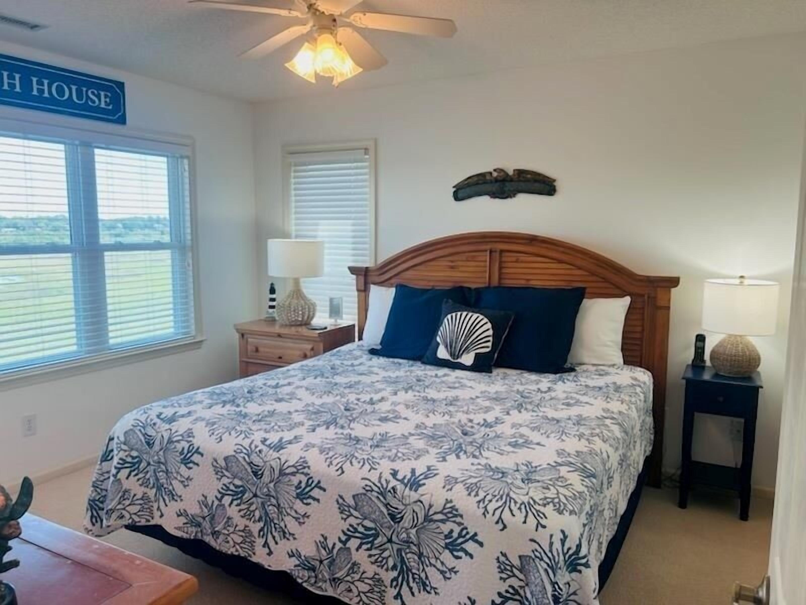 Une chambre à coucher comprenant un lit, deux tables de chevet, un ventilateur de plafond et une fenêtre avec des stores.