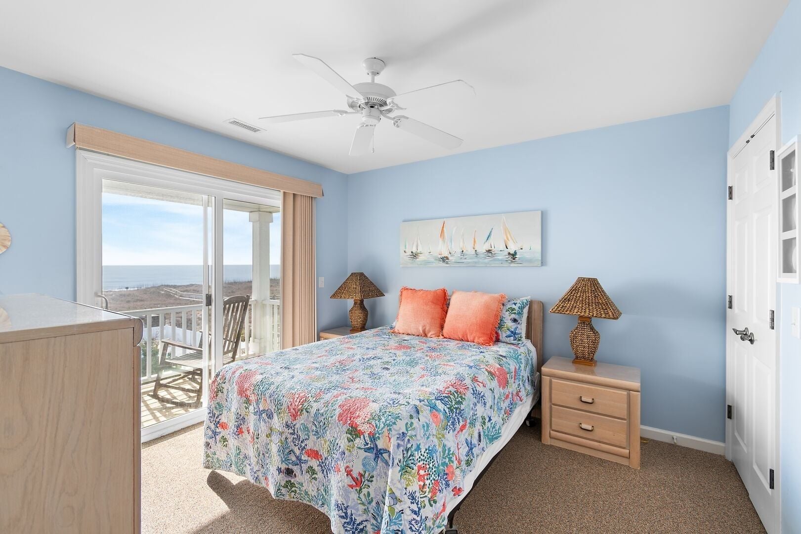 A bedroom with a bed, two nightstands, a ceiling fan, a painting, and a view of the ocean.
