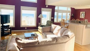 A living room with a beige sofa, a television, and a view of the beach through large windows. - License To Chill 4 Bedroom Home by RedAwning (Supply)